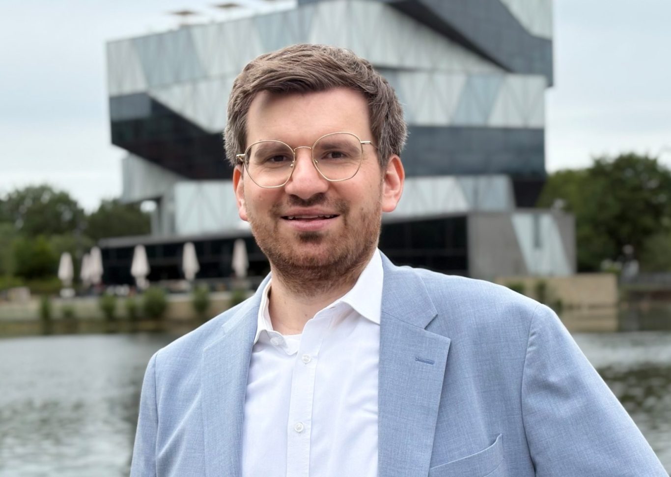 Rechtsanwalt Domenico Burkart Rechtsanwalt Burkart in hellblauem Jackett steht lächelnd vor Experimenta-Gebäude und Neckar.